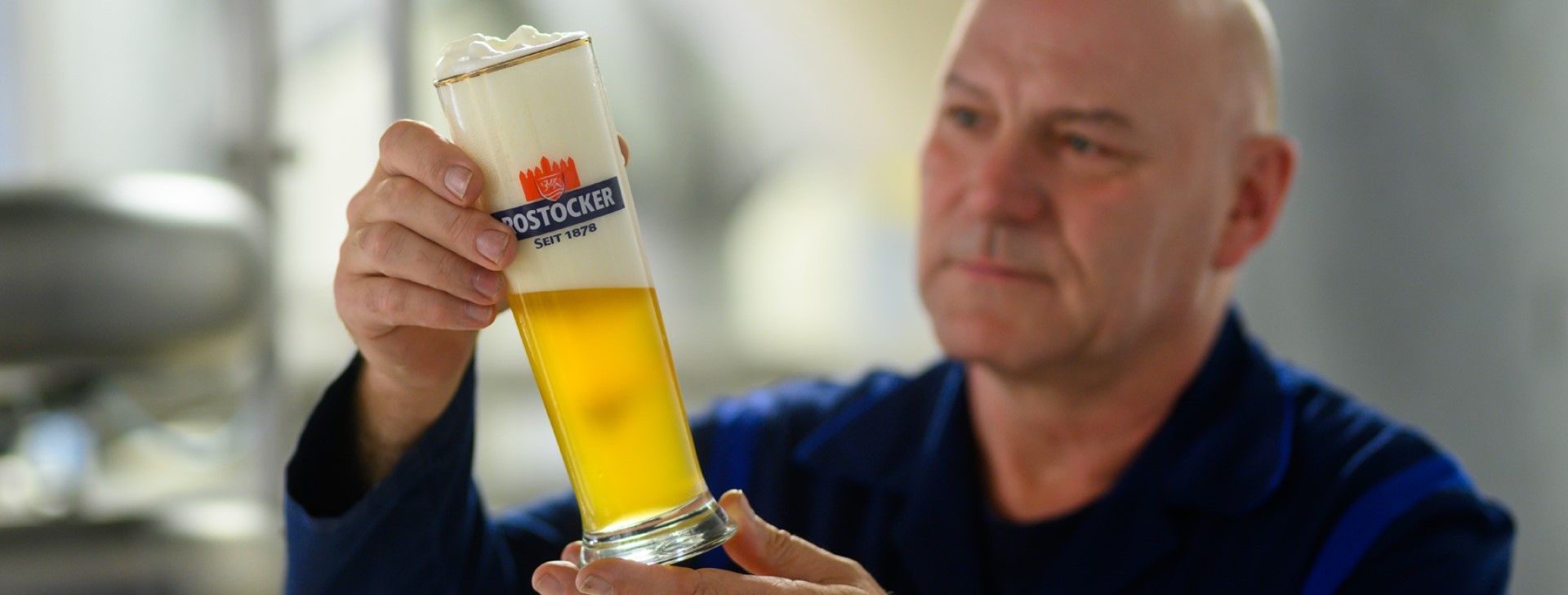 Ein älterer Mann in Arbeitskleidung prüft ein helles Bier in einem Glas.