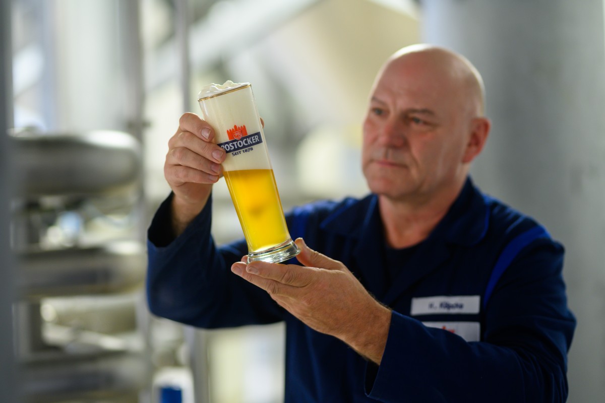 Ein Mann in einer blauen Arbeitskleidung prüft konzentriert ein Glas Bier.