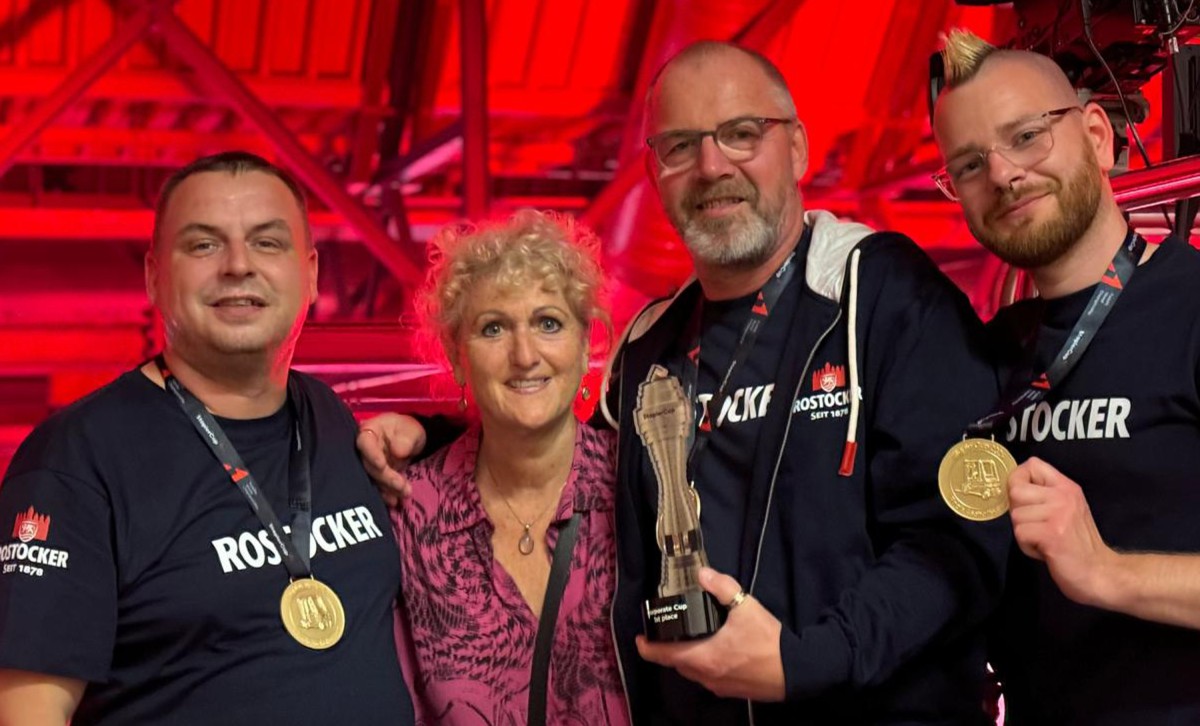Vier Personen posieren lächelnd mit Medaillen und einem Pokal vor einer roten Bühnenbeleuchtung.
