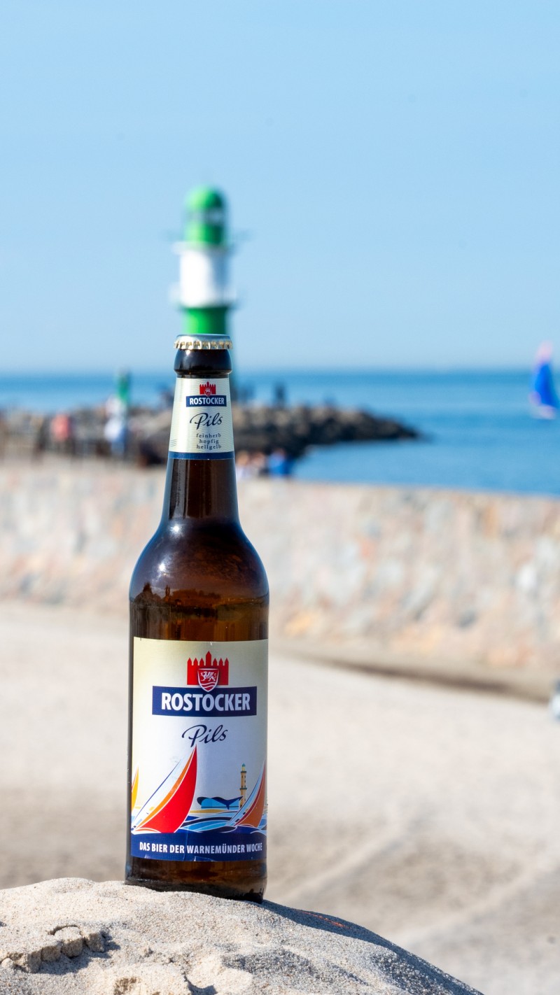 Ein Flasche Rostocker Pils, mit einem Segelboot auf dem Etikett, steht auf einem Stein, dahinter Meer.