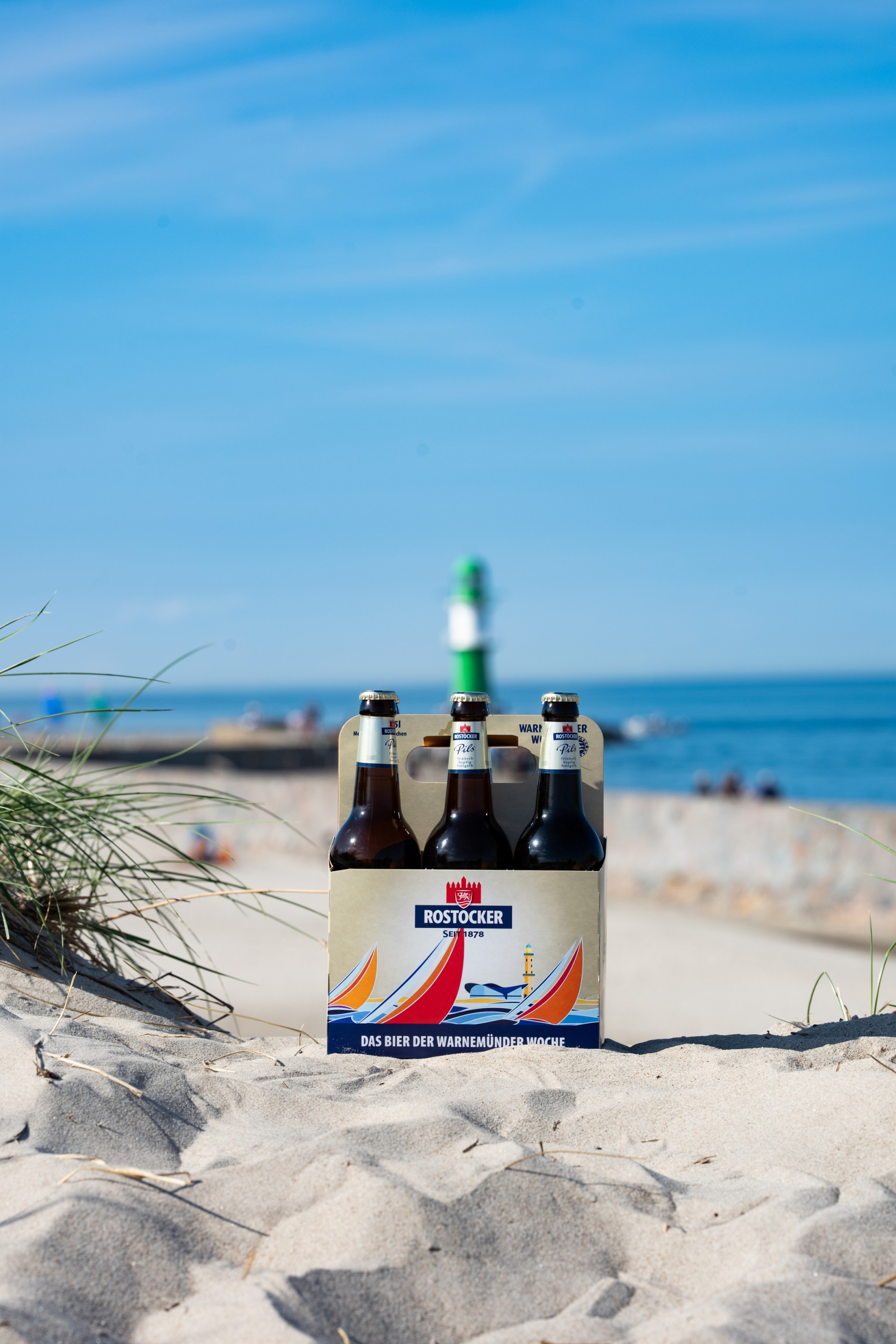 Ein Rostocker Pils Sechser Träger steht im Sand, dahinter ein grün weißer Leuchtturm.