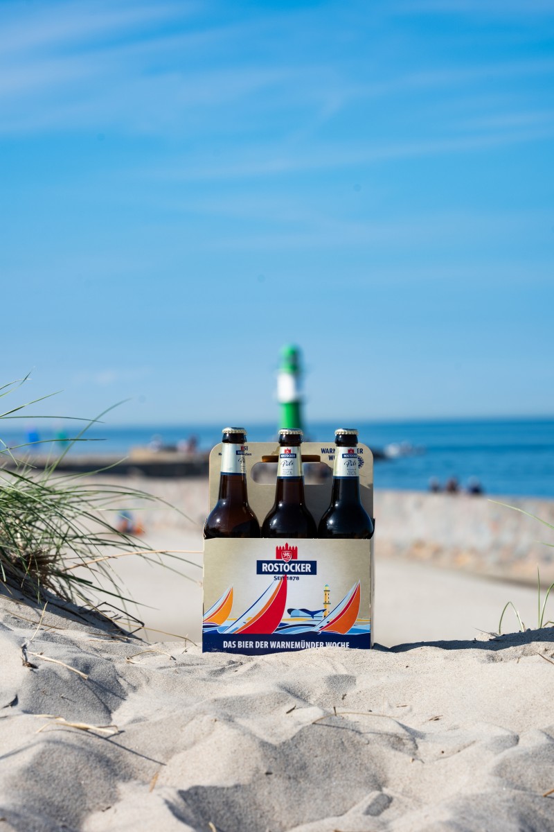 Ein Rostocker Pils Sechser Träger steht im Sand, dahinter ein grün weißer Leuchtturm.