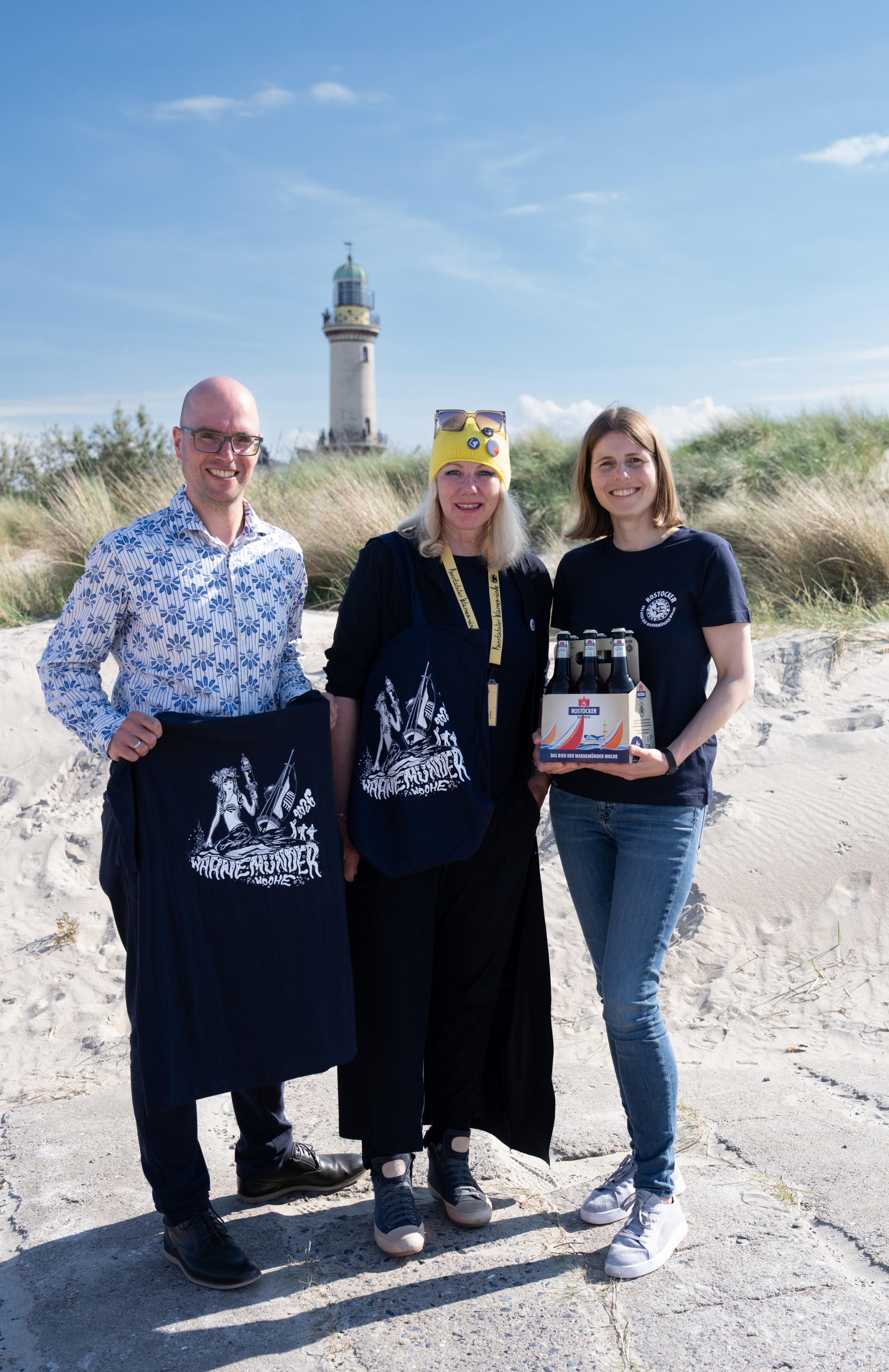 Drei Personen mit verschiedenen Rostocker Pils Artikeln stehen vor einer Dühne, dahinter ein Leuchtturm.
