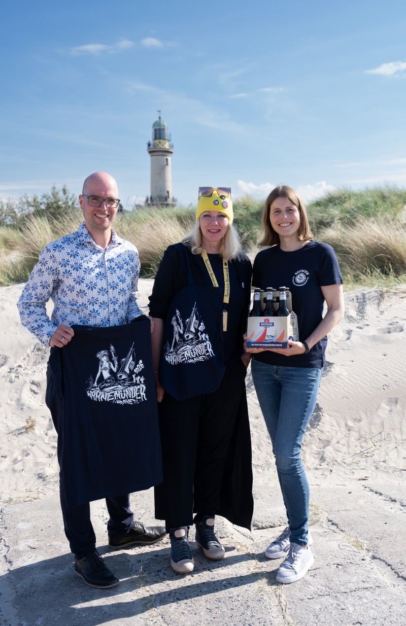 Drei Personen mit verschiedenen Rostocker Pils Artikeln stehen vor einer Dühne, dahinter ein Leuchtturm.