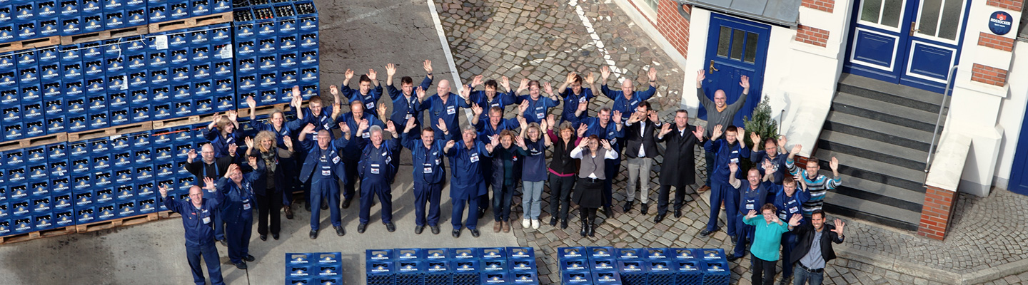 Mehrere Menschen winken freundlich auf einem Fabrikgelände neben gestapelten blauen Kisten.